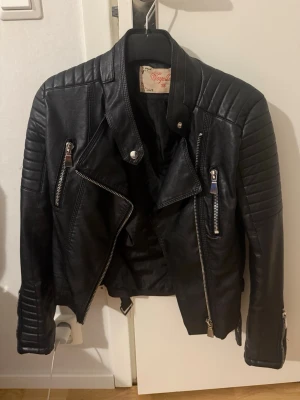Svart biker skinnjacka Viyella - Snygg svart skinnjacka från Viyella i klassisk bikerstil med dragkedjor framtill och på ärmarna. Jackan har vadderade detaljer på axlar och armar samt snedställda fickor med dragkedja. Perfekt för en edgy och cool look.