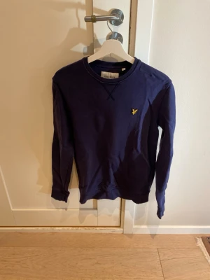 Mörkblå sweatshirt från Lyle & Scott - Mörkblå sweatshirt från Lyle & Scott med klassisk rund halsringning och gul logobrodyr på bröstet. Tröjan har långa ärmar och ribbade muddar vid ärmslut och nederkant. Perfekt för en avslappnad och stilren look.