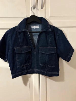 Mörkblå croppad jeansjacka Zara - Snygg croppad jeansjacka från Zara i mörkblå denim. Jackan har korta ärmar, två stora bröstfickor med lock och markerade sömmar för en cool look. Perfekt till ett par höga jeans eller kjol. Materialet är klassisk denim i bomull.