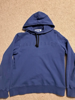 Blå hoodie från Jacob Cohën - Snygg blå hoodie från Jacob Cohën med broderad logga på bröstet. Hoodien har svarta dragsnören och en klassisk huva. Tillverkad i mjukt bomullsmaterial och har ribbade muddar vid ärmslut och nederkant. Perfekt för en avslappnad och trendig look.