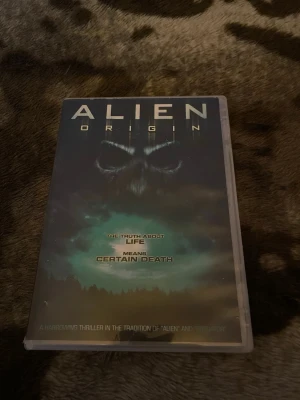 Alien Origin DVD thriller - DVD-film med titeln 'Alien Origin'. Omslaget är mörkt med ett mystiskt ansikte och gröna nyanser. Filmen är en thriller och har åldersgräns 15 år. Text på engelska och flera språk på baksidan. Perfekt för dig som gillar sci-fi och spänning.