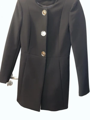 Svart kappa med stora knappar - Stilren svart kappa med raka linjer och tre stora, blanka knappar framtill. Kappan har en klassisk, enkel design med rund halsringning och långa ärmar. Perfekt för dig som gillar minimalistisk stil.