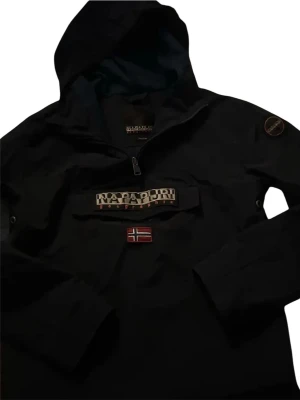 Svart vindjacka från Napapijri - Svart vindjacka från Napapijri med huva och dragkedja vid halsen. Stor logotyp på bröstet och norsk flagga framtill. Jackan har en klassisk anorak-design och är tillverkad i slitstarkt material, perfekt för blåsiga dagar.