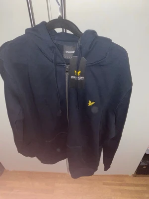 Svart hoodie från Lyle & Scott - Snygg mörkblå zip hoodie från Lyle & Scott med gul logga på bröstet. Klassisk modell med dragkedja, huva och fickor framtill. Perfekt för en avslappnad och stilren look. Materialet är mjukt och bekvämt, passar till det mesta.