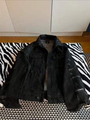 Dior strip jacket - Säljer en svart jeansjacka från Dior med distressed detaljer och slitningar för en edgy look. Klassisk modell med två bröstfickor och knappar framtill. Perfekt för dig som vill ha en trendig och exklusiv jacka i garderoben.