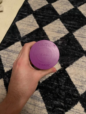 Lila timglas från TickiT 15 min - Säljer ett lila timglas från TickiT som mäter 15 minuter. Timglaset har en cylinderformad design med genomskinlig kropp och lila lock och botten. Perfekt som timer eller dekorativ detalj.