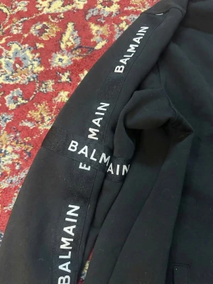 Svart zip sweater från Balmain - Snygg svart zip sweater från Balmain med dragkedja framtill och vita Balmain-loggor längs båda ärmarna. Tröjan har ribbade muddar och två fickor framtill. Tillverkad i mjukt bomullsmaterial som är skönt att bära. Hög kvalitet