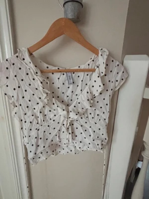 Vit prickig croppad blus från MNG - Supersöt vit croppad blus från MNG med svarta prickar, volangkant vid v-ringningen och knyt framtill. Blusen har korta ärmar och är tillverkad i ett lätt och luftigt material, perfekt för varma dagar.