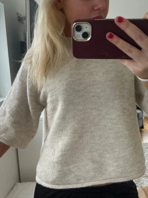 Beige stickad tröja med kort ärm - Mysig beige stickad tröja med korta ärmar och rund hals. Tröjan har en rak passform och är tillverkad i ett mjukt material som känns skönt mot huden. Perfekt för lager-på-lager och enkel att matcha med olika outfits.