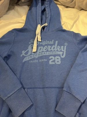 Blå Superdry hoodie med tryck - Snygg blå hoodie från Superdry med stort tryck på bröstet och texten 'The Original Superdry International 28'. Hoodien har huva med vita snören och en klassisk känguruficka framtill. Tillverkad i mjukt bomullsmaterial och har ribbade muddar vid ärmar och nederkant. Storlek M men passar bättre på en liten S hörte bra skick