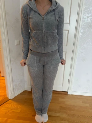 Juicy Couture Grå zip-hoodie med huva - Mjuk och skön grå hoodie med dragkedja och huva. Tröjan har ribbade muddar, fickor framtill och snören i huvan. Perfekt för chill dagar och enkel att matcha med jeans eller mjukisbyxor.
