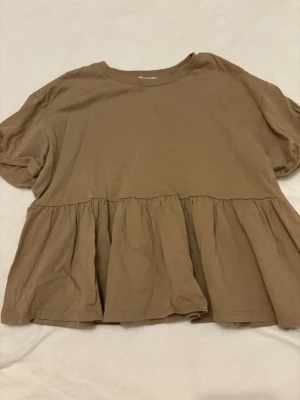 Beige volangtop från Bershka - Säljer en beige topp från Bershka med volang nedtill och rund halsringning. Toppen har korta ärmar och en loose passform som ger en avslappnad look. Perfekt att styla med jeans eller kjol för en trendig outfit.