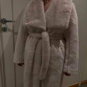 Ljusbeige fuskpäls kappa med skärp - Snygg ljusbeige kappa i fuskpäls med bred krage och knytband i midjan. Kappan har en lyxig känsla och stängs med både knappar och skärp. Perfekt för dig som vill ha en trendig och varm look under kalla dagar.