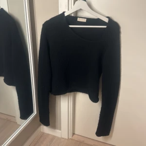 Svart stickad tröja med asymmetrisk hals - Trendig svart stickad tröja med lång ärm och en cool asymmetrisk halsringning. Modellen är croppad och har en mjuk, lite luddig struktur. Perfekt att styla med höga jeans eller kjol för en avslappnad look. Storlekstår ej men passar xxs-s - ingen defekt men lite nopprig.