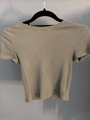 Beige ribbad croppad topp - Snygg beige croppad topp med ribbad struktur och korta ärmar. Perfekt till höga jeans eller kjol för en trendig look. Mjuk och stretchig, sitter tajt och bekvämt.