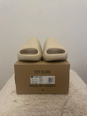 Bone 2022-2023 Yeezy Slide från Adidas - Säljer ett par Yeezy Slide från Adidas i färgen beige. Slidesen har en chunky, minimalistisk design med öppen tå och räfflad sula för extra grepp. Tillverkade i ett mjukt och flexibelt syntetmaterial som ger skön komfort. Perfekta för en avslappnad och trendig look.