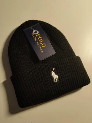 Svart ribbstickad mössa Polo Ralph Lauren - Stilren svart ribbstickad mössa från Polo Ralph Lauren med klassisk vit broderad logga framtill. Mössan har uppvikt kant och är tillverkad i mjukt material som håller värmen. Perfekt accessoar för kalla dagar och enkel att matcha med olika outfits