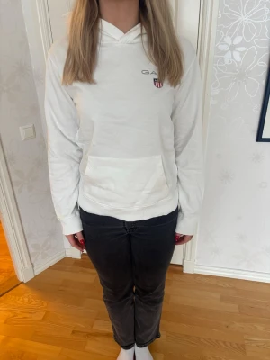 Vit hoodie från Gant med logga - Säljer en stilren vit hoodie från Gant med liten broderad logga och sköld på bröstet. Hoodien har huva, känguruficka och ribbade muddar vid ärmslut och nederkant. Perfekt för dig som gillar klassisk och enkel stil. Storleken motsvarar M