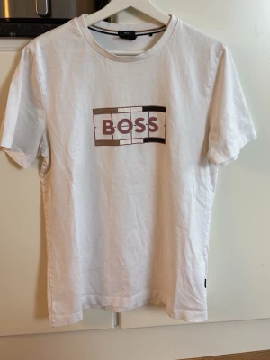 Vit BOSS t-shirt med tryck - Snygg vit t-shirt från BOSS med ett stort tryck på bröstet där det står 'BOSS' i brunt och svart. Klassisk passform och rund halsringning. Tillverkad i mjuk bomull som känns skön mot huden. Perfekt för en clean och stilren look.