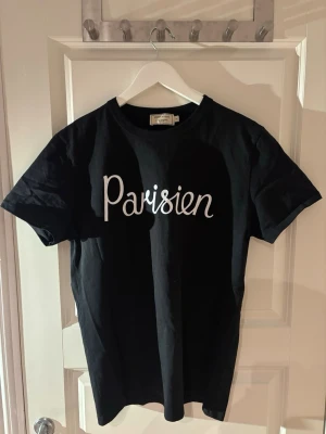 Svart Parisien t-shirt Maison Kitsuné - Snygg svart t-shirt från Maison Kitsuné med vit text 'Parisien' på bröstet. Klassisk rund halsringning och korta ärmar. Tillverkad i mjuk bomull som känns skön mot huden. Perfekt för en avslappnad och trendig look.