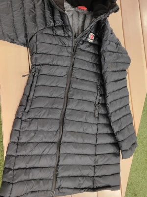 Svart lång dunjacka från Fjällräven - Snygg svart lång dunjacka från Fjällräven med broderad logotyp på bröstet. Jackan har horisontella sömmar, dragkedja framtill och två sidofickor med dragkedja. Perfekt för kalla dagar och riktigt skön att bära.