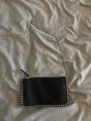 Svart kuvertväska med silverkedja - Snygg svart kuvertväska med silverfärgade nitar längs kanterna och en lång silverkedja som axelrem. Väskan har en enkel, rektangulär form och stängs med dragkedja upptill. Perfekt för dig som gillar en edgy och stilren look.