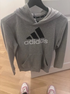 Grå hoodie från Adidas med logga - Snygg grå hoodie från Adidas med stor svart och vit logga på bröstet. Klassisk känguruficka framtill och huva. Tillverkad i mjukt bomullsmaterial som är skönt att ha på sig. Perfekt för en avslappnad och sportig stil.