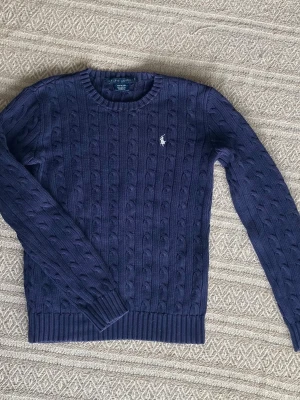 Mörkblå kabelstickad tröja Ralph Lauren - Kabelstickad tröja i mörkblått från Ralph Lauren med klassisk rund halsringning och vit broderad logga på bröstet. Tillverkad i 100% bomull och har ribbade muddar vid ärmslut och nederkant. Perfekt för en stilren och tidlös look. OBS defekt att taggen sitter löst som syns på bild med går att fixa lätt om man vill