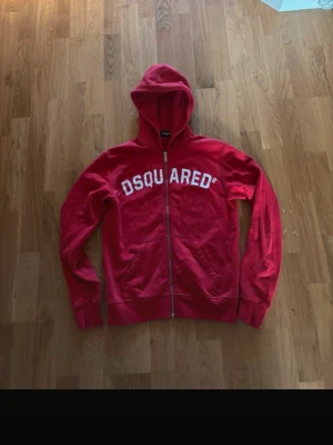 Röd hoodie från Dsquared2 med dragkedja - Röd hoodie från Dsquared2 med vit logga tryckt över bröstet. Modellen har huva, hel dragkedja framtill och två fickor. Perfekt för en avslappnad och trendig stil. Är slimfit och är i barn storlek. 