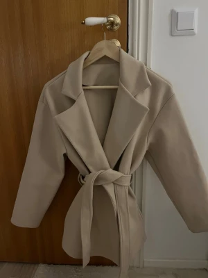 Beige kappa med knytskärp - Stilren beige kappa med bred krage och knytskärp i midjan. Kappan har en rak passform och är tillverkad i ett mjukt tyg som ger en clean look. Perfekt för dig som gillar minimalistisk och trendig stil. Tyvärr två små fläckar på insidan av kappan, inget som påverkar passform eller utseende på.