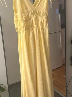 Gul långklänning med knytband - Säljer en ljusgul långklänning med v-ringning och smala axelband som knyts på axlarna. Klänningen har resår i midjan för en bekväm passform och ett mjukt, luftigt tyg som passar perfekt till varma dagar.