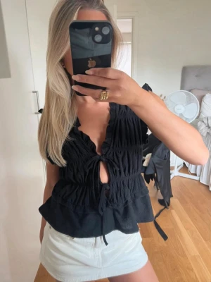 Svart topp från gina tricot - Trendig svart omlottopp med knyt framtill och volangkant. Toppen är ärmlös och har en djup v-ringning. Tillverkad i ett lätt, strukturerat material som ger en luftig känsla. Perfekt att styla med kjol eller jeans.
