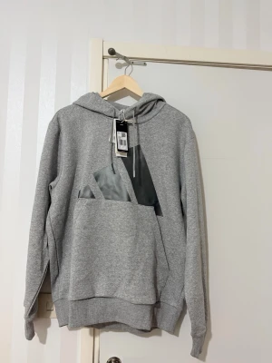 Grå hoodie från Adidas med logga - Snygg grå hoodie från Adidas med stor svart logga på bröstet. Klassisk känguruficka framtill, ribbade muddar och justerbar huva med dragsko. Tillverkad i mjukt sweatshirtmaterial som passar perfekt till en avslappnad stil.
