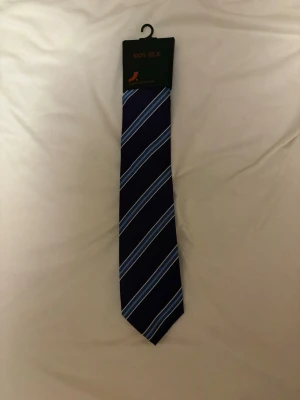 Mörkblå sidenslips med ränder - Snygg mörkblå slips i 100% siden med diagonala ränder i ljusblått och vitt. Perfekt accessoar för att lyfta din outfit med en klassisk och stilren touch. Slipsen har en elegant glans och känns lyxig.