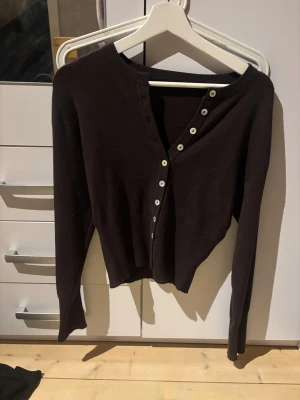 Mörkbrun kofta med knappar - Snygg mörkbrun kofta med långa ärmar och vita knappar framtill. Koftan har en klassisk, rak passform och ribbstickade muddar vid ärmslut och nederkant. Perfekt att slänga över en t-shirt eller linne för en chill look.