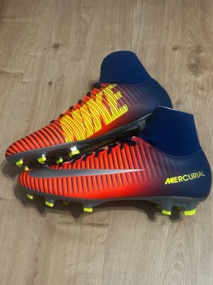 Nike Mercurial röd/blå fotbollsskor - Snygga Nike Mercurial fotbollsskor i rött och blått med gula detaljer och högt skaft. Skorna har en strumpliknande passform, gula dobbar och stora Nike-loggor på sidorna. Perfekta för dig som vill sticka ut på planen och få bra grepp på gräs.
