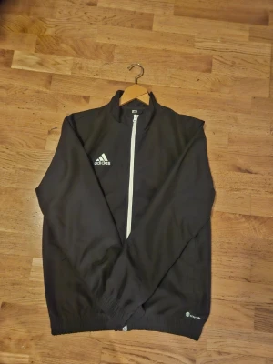 Svart Adidas träningsjacka - Snygg svart träningsjacka från Adidas med vit dragkedja och logga på bröstet. Jackan har ståkrage, elastiska muddar vid ärmslut och nederkant samt två sidofickor. Tillverkad i ett lätt och funktionellt material, perfekt för träning eller sport.