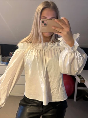 Vit offshoulder blus med puffärmar - Trendig vit offshoulder blus med långa puffärmar och smockad resår vid axlarna. Ärmarna har dekorativa knytband och små gulddetaljer vid ärmsluten. Tillverkad i ett lätt och luftigt tyg, perfekt för en stilren look.