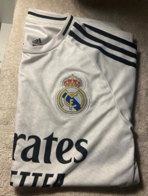 Real Madrid matchtröja Adidas vit - Säljer en vit Real Madrid matchtröja från Adidas med klubbmärke på bröstet och svarta detaljer. Tröjan har svarta ränder på axlarna och trycket 'Emirates Fly Better' på framsidan. Perfekt för fotbollsfans som vill stötta sitt lag.