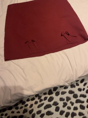 Vinröd kjol med rosetter - Säljer en vinröd kjol i kort modell med två söta rosetter framtill. Kjolen har en enkel, rak passform och är tillverkad i ett mjukt tyg som känns bekvämt. Perfekt att matcha med en stickad tröja eller crop top.