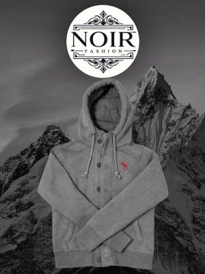 Grå hoodie från Abercrombie & Fitch - Snygg grå hoodie från Abercrombie & Fitch med knappar framtill och dragsko i huvan. Har en rosa logga på bröstet och mjukt, quiltat innerfoder i huvan. Perfekt för dig som gillar en avslappnad och trendig stil.