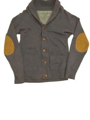 Kofta - Snygg grå kofta med stora bruna armbågslappar och träknappar framtill. Koftan har två fickor fram och en bred krage som ger en chill look. Perfekt för lager på lager och passar till det mesta. Materialet känns mjukt och bekvämt. Storlek s