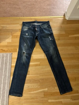 Mörkblå  jeans från Dsquared2 - Säljer ett par mörkblå jeans från Dsquared2 med slitningar och hål på flera ställen. Jeansen har raka ben, klassisk femficksmodell och detaljer som ger en cool, använd look. Perfekta för dig som gillar streetstyle och vill sticka ut.