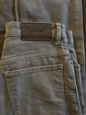 Beige/grå jeans från Just Junkies - Snygga beige/grå jeans från Just Junkies med klassisk femficksdesign och rak passform. Jeansen har en mörk läderpatch med logga bak i midjan och är gjorda i ett slitstarkt bomullstyg. Perfekta för en avslappnad och trendig look.