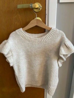 Beige stickad topp med volangärm - Jättesöt stickad topp med volang ärm. Den är ifrån Ginatricot Young i storlek 146/152 men den är ganska kort i modellen. Den är alldrig andvänd.