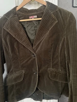 Brun manchesterkavaj från Cue - Snygg brun grön  kavaj i manchestertyg från Cue med tunna vita ränder. Klassisk krage, två knappar framtill och fickor med lock. Figursydd modell som ger en schysst siluett. Perfekt för dig som vill ha en retrovibe i garderoben.