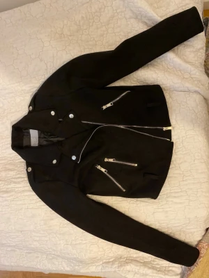 Svart skinnjacka med silvriga dragkedjor - Snygg svart skinnjacka i klassisk bikerstil med flera silvriga dragkedjor framtill och tryckknappar vid kragen. Jackan har figurnära passform och långa ärmar. Perfekt för dig som vill ha en edgy och stilren look.