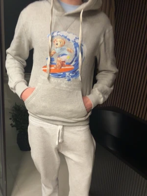 Grå hoodie Polo Ralph Lauren - Säljer en ljusgrå hoodie från Polo Ralph Lauren med en färgglad print av en björn som surfar på bröstet. Hoodien har huva med dragsko och en stor magficka. Tillverkad i mjukt bomullsmaterial, perfekt för chill dagar.