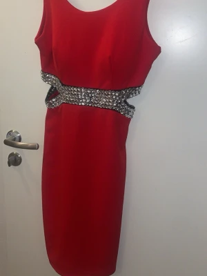 Röd klänning med paljettbälte - Snygg röd klänning i figurnära modell med bredt paljettbälte i midjan som ger extra bling. Ärmlös design och rund halsringning. Perfekt för dig som vill sticka ut med en elegant och glittrig look.