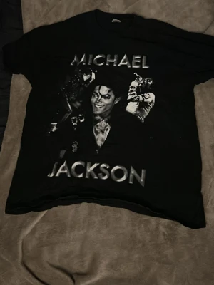 Svart Michael Jackson t-shirt - Cool svart t-shirt med stort Michael Jackson-tryck på framsidan och tryck med signatur och datum på baksidan. T-shirten har korta ärmar och är i klassisk passform. Perfekt för dig som gillar musik och vill sticka ut med en ikonisk look. Stor storlek men har själv medium annars i storlek så passar fint som oversized